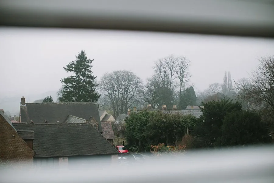 Misty view through window blinds of tiled suburban rooftops and bare trees on a wet grey day