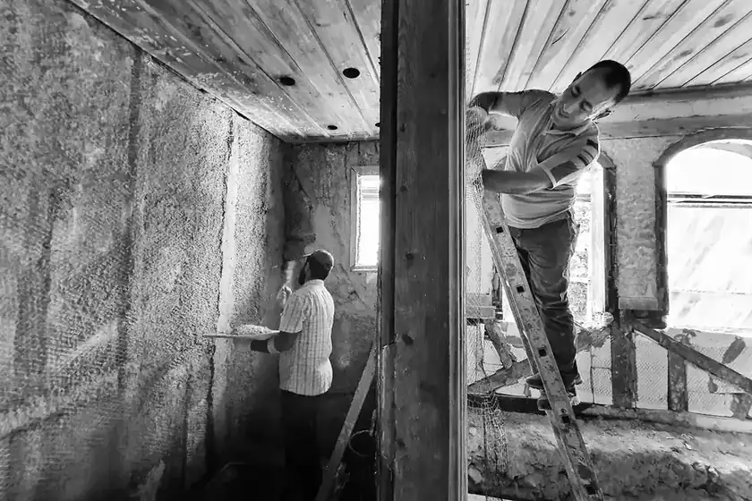 Black and white photo of two workers renovating a room, one plastering a wall and one on a ladder at the ceiling