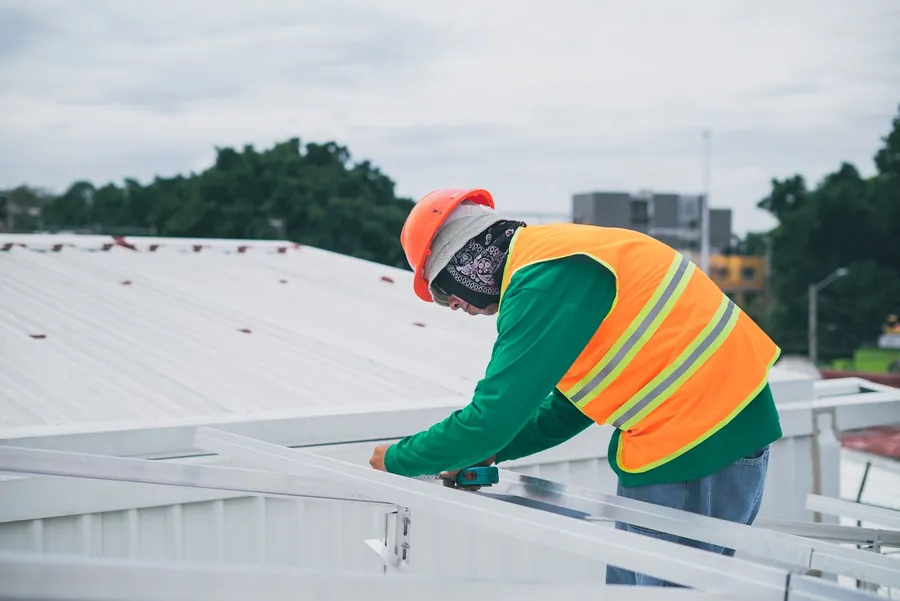 Roofer in a hard hat and high-visibility jacket working on a pale metal roof