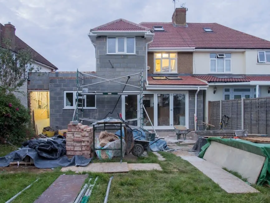 Rear extension of a semi-detached house under construction, with block walls, a temporary flat roof and building materials in the garden